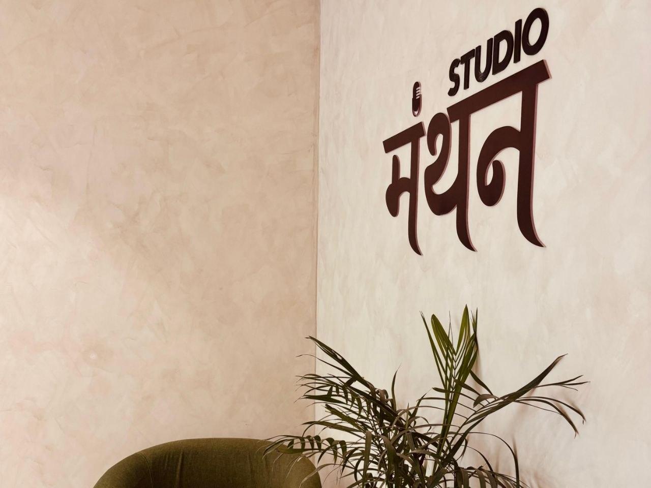 Interior of Manthan Studio in Hauz Khas — warm wood paneling, a vintage microphone on a boom arm, and trailing plants near a window.